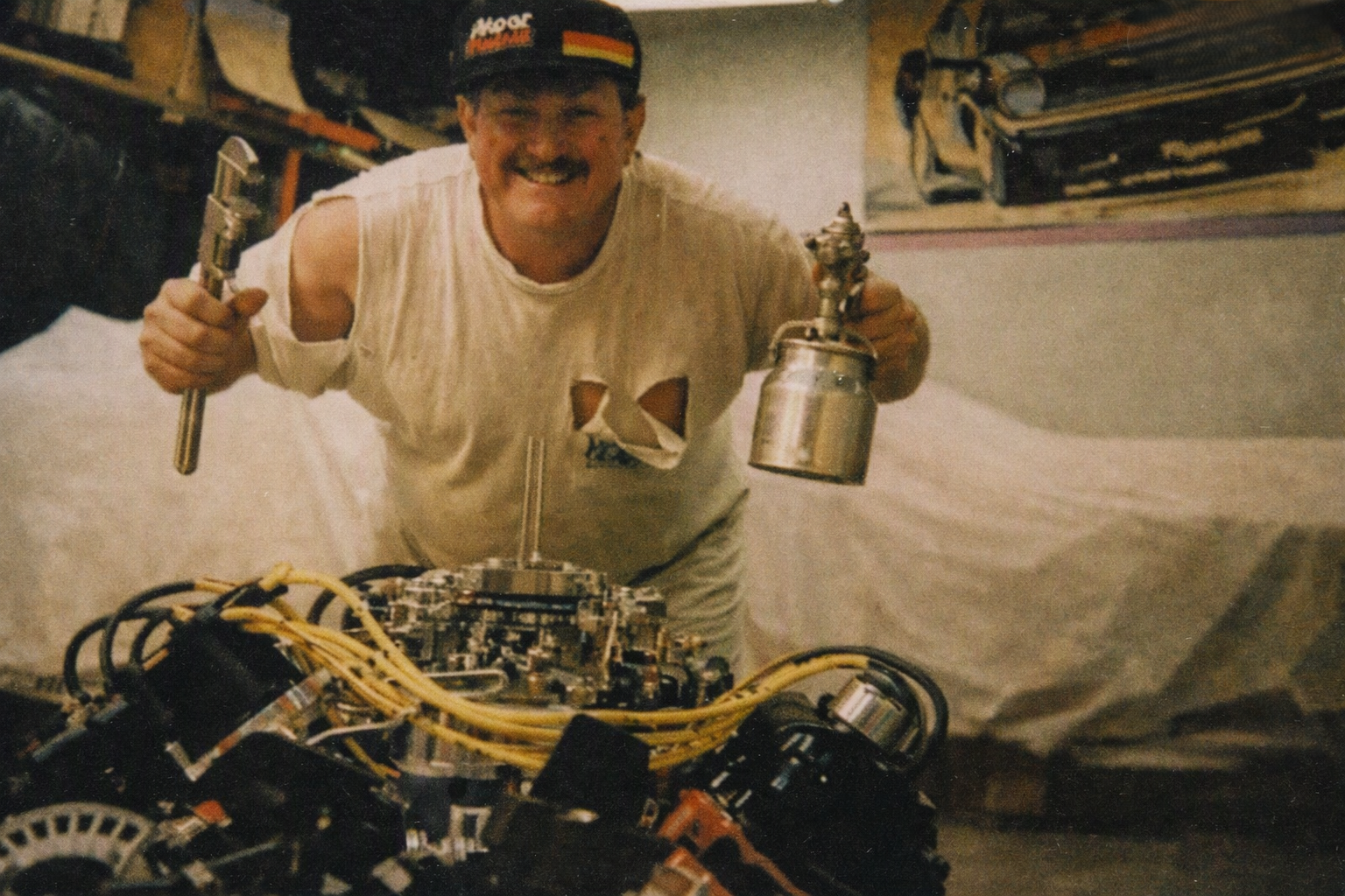 Dan Powell in his Collinsville Oklahoma shop — grinning over a freshly built 426 Hemi engine, wrench in one hand, spray gun in the other